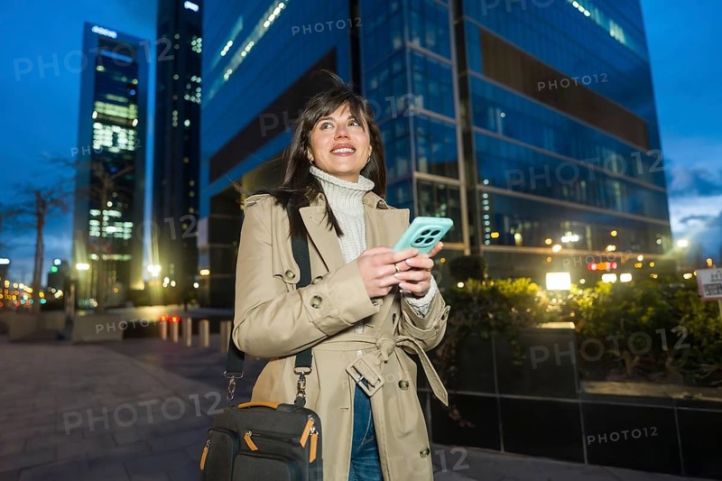 Young professional woman walking along a city sidewalk at dusk, holding a smartphone and carrying a laptop bag, navigating the urban environment between modern high rise buildings