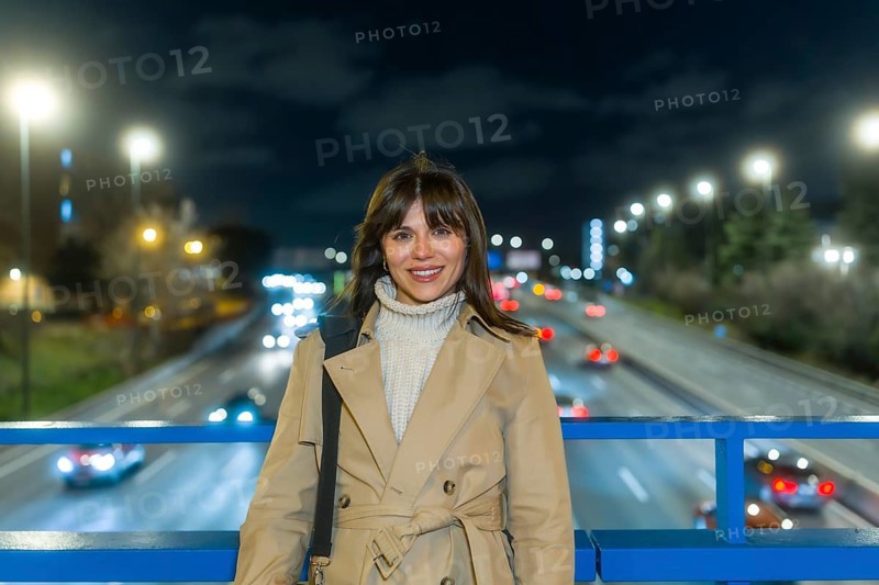 Woman smiling and standing on a bridge over a blurred highway with car lights creating bokeh, reflecting urban movement, modern city life, and evening commute