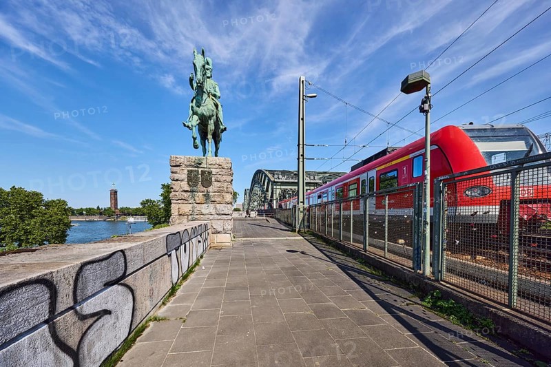 Equestrian statue of Emperor Frederick III, Hohenzollern Bridge, arched bridge, steel arches, metal fence, sandstone wall, S-Bahn, public transport, overhead lines, lantern, graffiti, footpath made of paving tiles, trees, blue sky, cirrus clouds, Cologne, district-free city, North Rhine-Westphalia, Germany