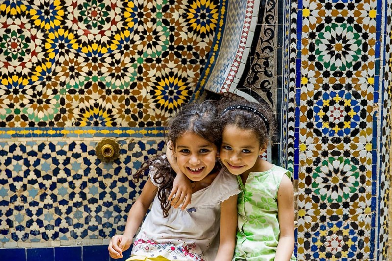 Two smiling girls in front of a wall with colorful tiles, alley in the old town, Souk, Fez El Bali, Medina, UNESCO World Heritage Site, Fez, Morocco