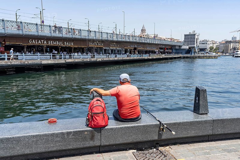 Istanbul, Turkey. June 23rd 2025. A mam sits fishing on the dockside in Eminonu beside the famous Galata Bridge with its fish restaurants and the iconic Galata Tower, Istanbul, Turkey