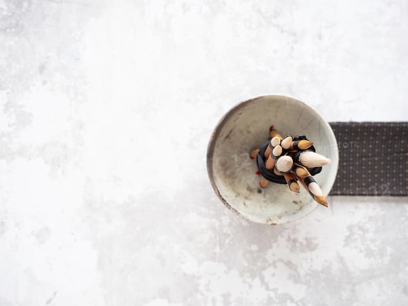 Paint brushes with bamboo handles are bundled together in a ceramic bowl, resting on a white table next to a patterned cloth