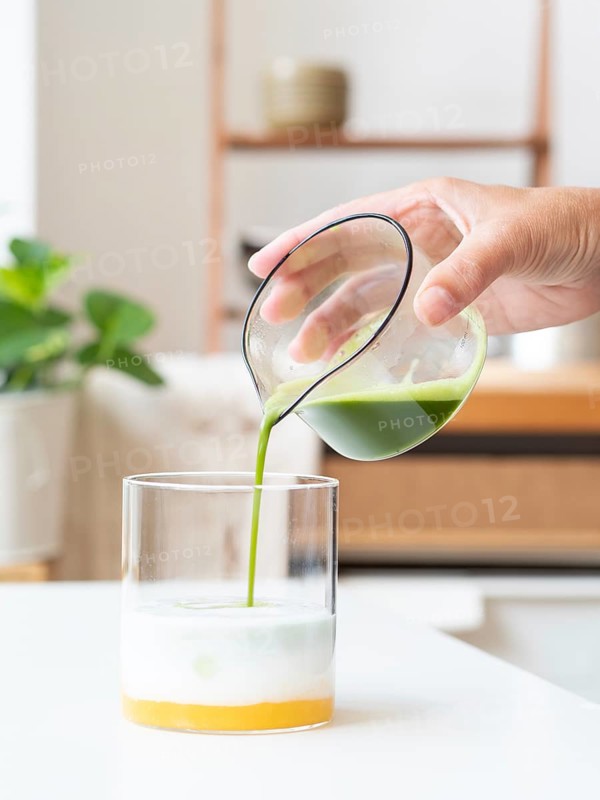 Hand pouring vibrant green matcha into a glass of mango base and milk, creating a layered drink