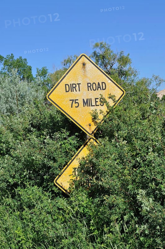 A yellow road sign with the words 'Dirt Road 75 Miles' appears from lush vegetation, Grand Staircase Escalante National Monument, Utah, USA