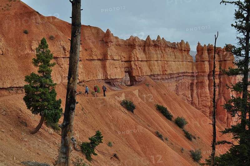 Hikers on a trail along dramatic red cliffs under looming clouds, Bryce Canyon National Park, Utah, USA