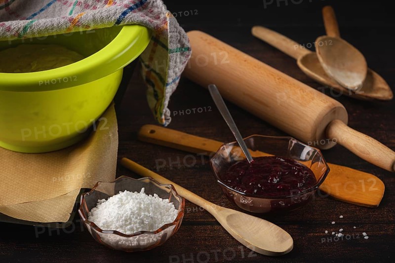 Dough bowl next to rolling pin, cooking spoon, baking paper, jam and sugar on a dark wooden table