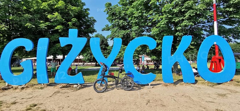 A bicycle stands in front of a large blue lettering of 'Gizycko' on a sandy beach, Masuria bicycle tour, Poland