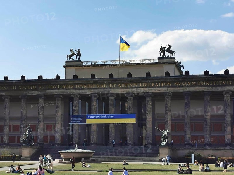 People relax on the lawn in front of the old museum on Museum Island in Berlin where the Ukrainian flag is blowing in the wind, Berlin