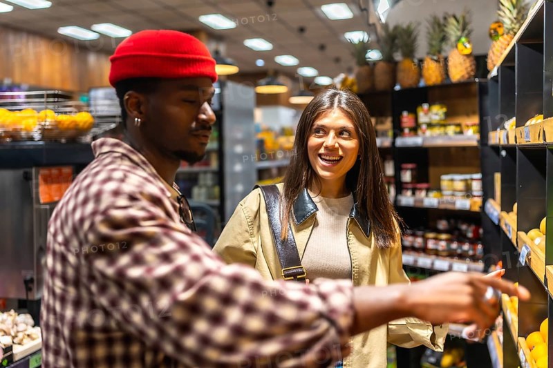 Young diverse couple shopping for food in a modern grocery store, with the man pointing to items on a shelf and the woman smiling happily during their shared activity