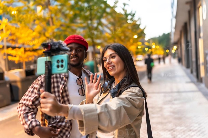 Diverse couple vlogging together on a city sidewalk, waving to camera held on a selfie stick, smiling as they stream and document their urban travel adventure for followers