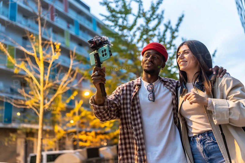 Diverse young couple vlogging urban exploration together, smiling and walking through the city while recording on a smartphone mounted to a gimbal for social media content and travel storytelling