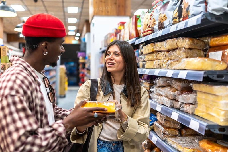 Diverse young couple looking at each other, one holding a packaged baked good while standing in a busy supermarket aisle, engaging in food choice and daily shopping