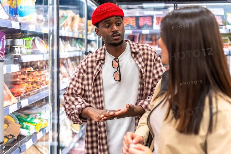 Black man in red cap and plaid shirt and white woman compare items in the refrigerated aisle while grocery shopping, discussing fresh food choices and planning purchases together