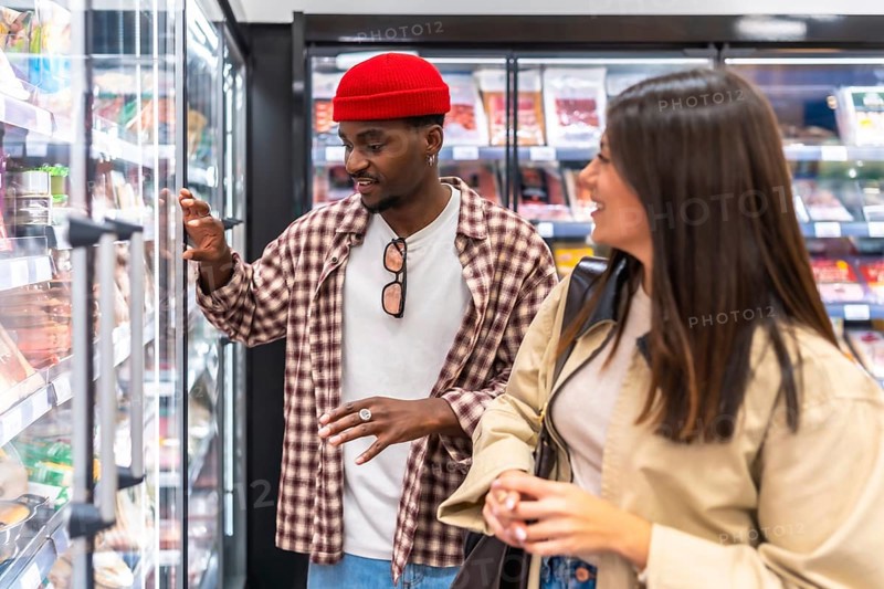Young diverse couple shopping for groceries at a modern supermarket, standing in front of refrigerated shelves and choosing fresh food items while talking and smiling