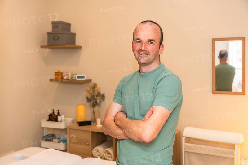 Professional man massage therapist smiling confidently, standing with crossed arms in a therapy room, ready to provide wellness and health services to clients