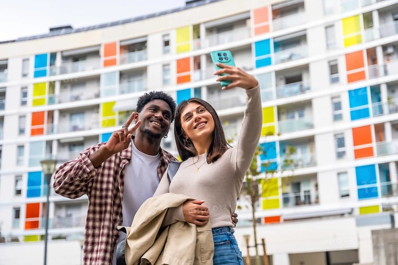 Happy diverse couple smiling and posing for a smartphone selfie, showing a peace sign while standing outdoors in front of a modern residential building with vibrant balconies