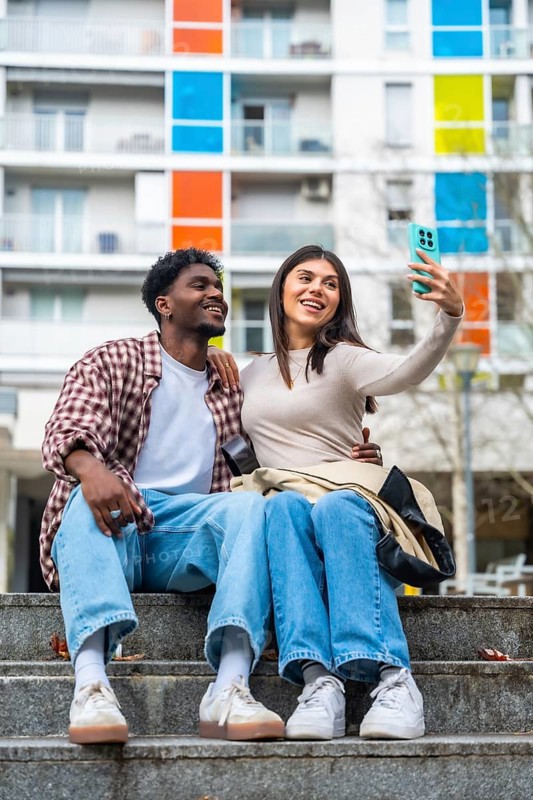 Happy diverse young couple sitting on concrete steps, smiling and taking a cheerful selfie together with a smartphone in a modern city setting with colorful buildings in the background
