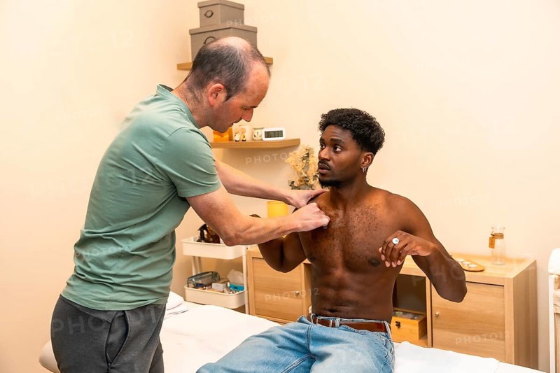 Physical therapist performing an examination and palpation on a young adult male patient's shoulder during a rehabilitation session in a professional clinic setting