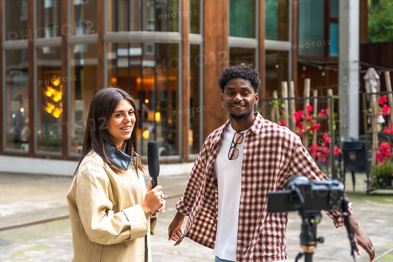 Journalist holding a microphone interviewing a young man on a city street, documenting the conversation with a camera on a tripod for a live broadcast or social media content
