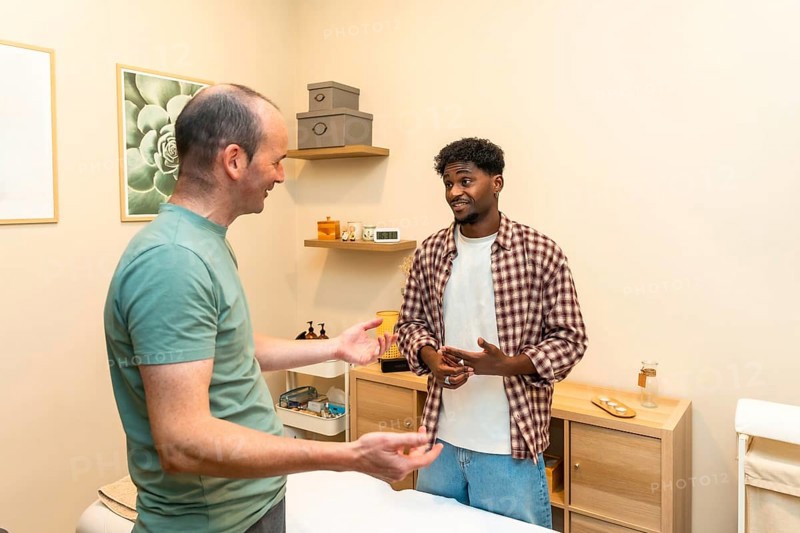 Two men engaging in a friendly and professional conversation about health and wellness, consulting with a specialist in a bright, modern clinic setting during their therapy appointment