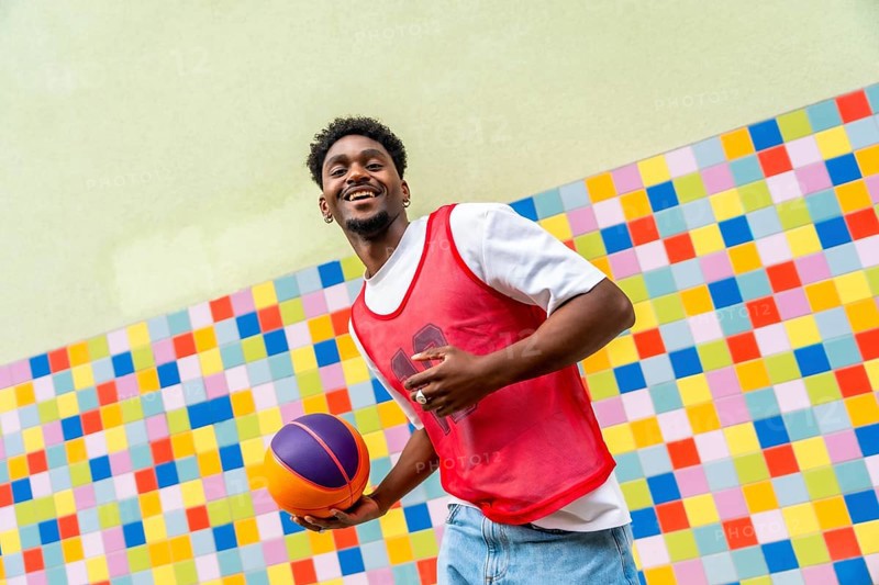 Young black man happily holding a basketball, dressed in a red sports vest and jeans, posing against a vibrant, mosaic tiled background, symbolizing urban youth and active lifestyle