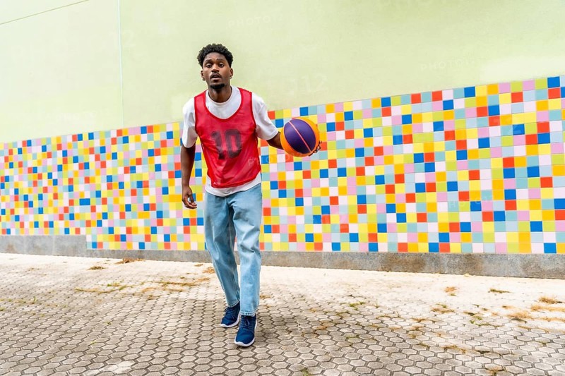 Young black man in casual sportswear playing and dribbling a basketball on a paved court against a vibrant, multi colored wall, representing youth, sport, and urban lifestyle