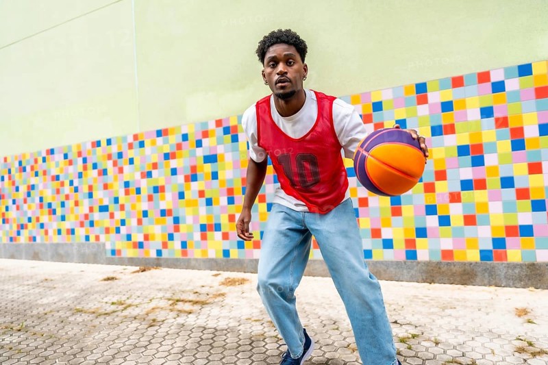 Young man actively playing street basketball, dribbling the ball with focus and energy against a vibrant, mosaic patterned wall, embodying urban sport and active lifestyle
