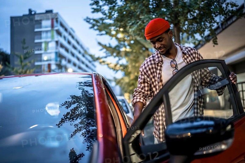 Young black man in a red beanie smiling as he opens a car door on a city street, ready to drive through an urban neighborhood with buildings and trees nearby