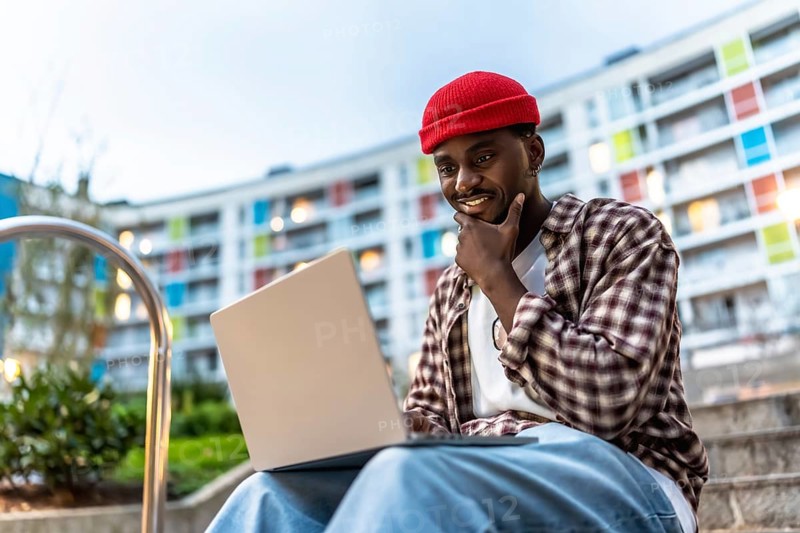 Young black man sitting on apartment steps with laptop, smiling and thoughtful as he works or studies outdoors, embodying modern urban remote work and digital lifestyle