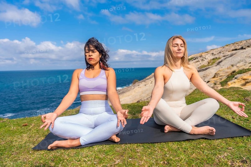 Two healthy multiracial women performing yoga and meditation poses in lotus position on a yoga mat during a mindful wellness activity outdoors by the ocean in nature