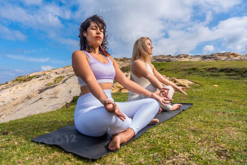 Two young women meditating on yoga mats outdoors, finding peace and mindfulness in nature, practicing spiritual wellness and healthy lifestyle under a blue sky