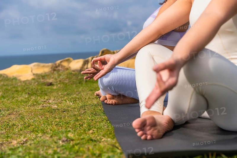 Two women practicing yoga and meditating outdoors on yoga mats, sitting in lotus pose with hands in jnana mudra, finding balance, wellness, and peace by the blue ocean and green grass