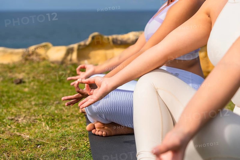 Women practicing meditation outdoors, sitting in a mudra position on yoga mats on a green cliffside, finding peace and spiritual balance overlooking the serene ocean