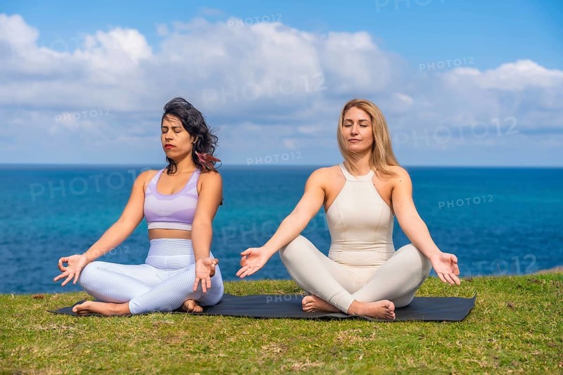 Two women sit cross legged on a mat by the ocean, practicing yoga and meditation together at sunrise, finding calm, balance and mindful connection with nature and each other