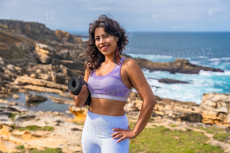 Woman standing on a rocky coastline holding a yoga mat, preparing for an outdoor fitness session with the ocean and sky creating a serene backdrop for health and wellness activities