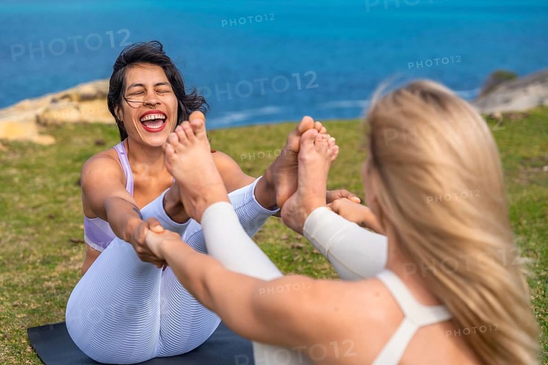 Two women performing a yoga pose together on a mat on green grass by the sea, connecting through shared laughter and exercise, promoting friendship and wellness in nature