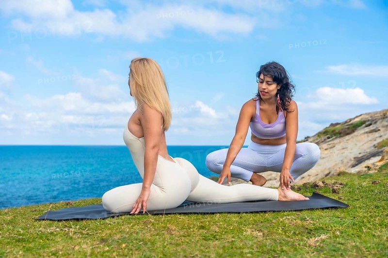 Two women are practicing yoga on a mat outdoors, with one instructor supporting a student in a stretching pose by the sea, highlighting wellness and healthy lifestyle