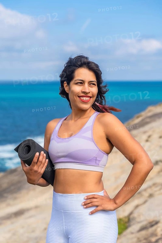 Woman holding a yoga mat and smiling, standing on a rocky cliff overlooking the blue ocean during daytime, enjoying an active and healthy lifestyle while practicing yoga and meditation in nature