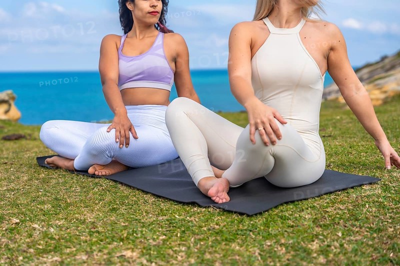 Two diverse women in seated spinal twist yoga poses on mats, meditating and connecting with nature by the ocean under a clear sky, calm and serene seaside wellness session