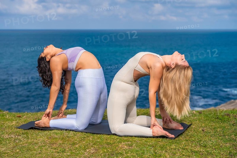 Two women performing ustrasana back to back on yoga mats, stretching their bodies and achieving flexibility during a meditation and wellness session by the ocean