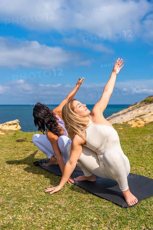 Two women practicing yoga on mats by a calm ocean, stretching into upward poses and finding balance, mindfulness and wellness under blue skies and gentle daylight