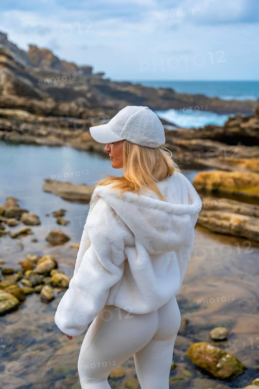 Woman in comfortable hoodie and leggings meditating on rocky coast, back to camera, finding calm and mindfulness beside the ocean under a tranquil sky, peaceful solitude