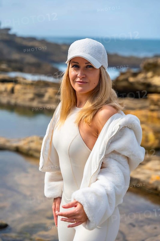 Woman in white sportswear and baseball cap stands on rocky shoreline by the blue sea, smiling confidently in sunny, relaxed outdoor fashion portrait evoking wellness and summer freedom