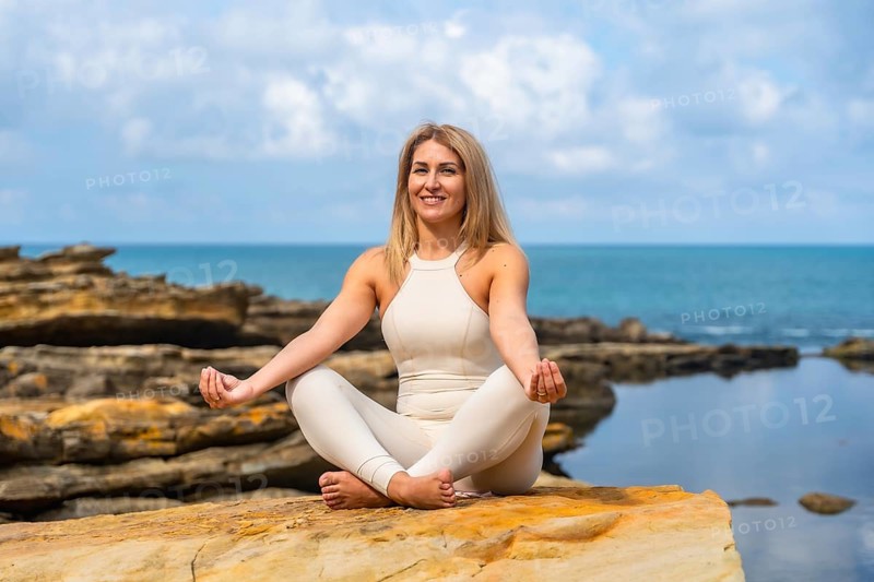 Woman practicing yoga in lotus on a rocky ocean cliff, meditating with eyes closed, embracing balance, calm and strength amid sea breeze and blue sky serenity