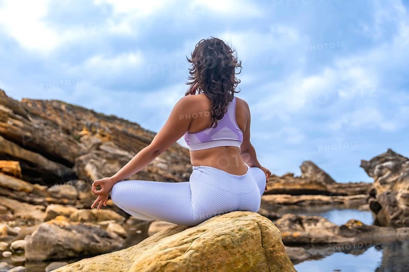 Woman sitting in a yoga lotus position on a rock, practicing meditation and mindfulness in a natural outdoor setting with rocky terrain and water pools under a cloudy sky