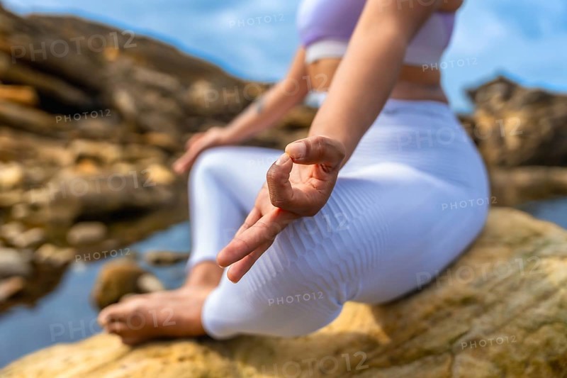 Woman performing yoga mudra hand gesture, finding inner peace and mindful wellness while sitting in nature on a rock with sky and water in the background