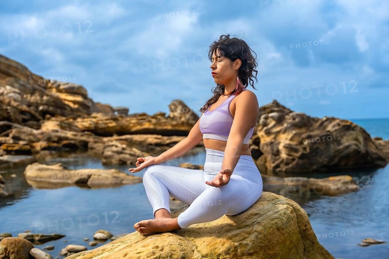 Woman sitting in a yoga pose, meditating outdoors on a rock by the sea, finding tranquility and balance amidst the natural coastal environment on a cloudy day