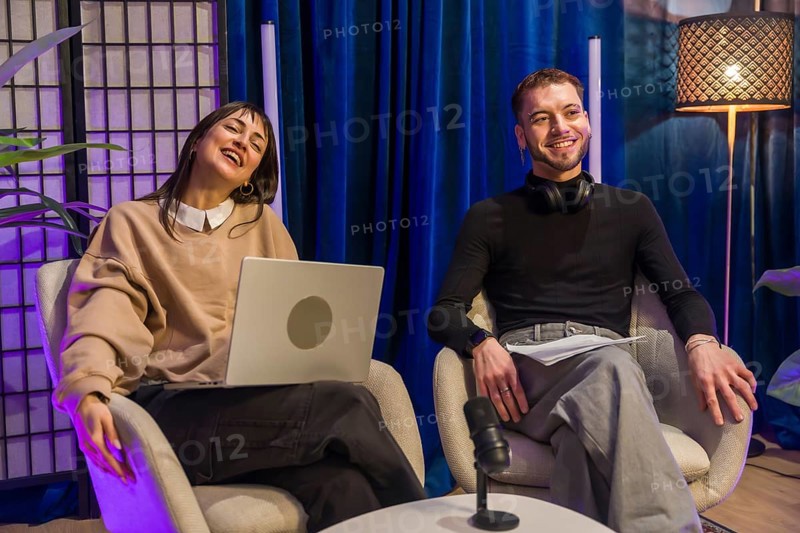 Two young diverse content creators smiling and sharing ideas during an online podcast recording session in a modern studio setup, capturing authentic moments for digital media