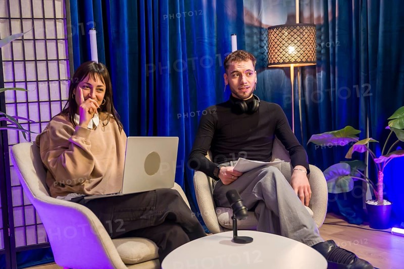 Two young content creators engaging in a podcast recording session, using a laptop, microphone, and headphones within a vibrant studio setup for social media and streaming