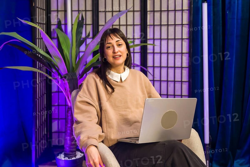 Woman sitting in a professional content creation studio, holding a laptop in her lap while looking towards the camera, ready for recording podcasts or social media videos